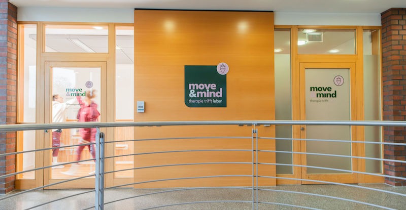 Aussenaufnahme-Praxisgebäude-move-and-mind-Hamburg-Harburg Außenaufnahme der Praxis Move & Mind in Harburg mit zwei Türen. Durch die erste Glastür sind verschwommen zwei Teammitglieder zu sehen, die sich "High Five" abklatschen