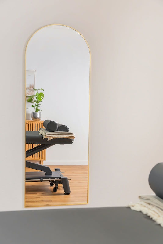 Behandlungszimmer-Praxis-Move-and-Mind-Harburg Blick in einen großen, torbogenförmigen Spiegel, worin ein Behandlungstisch mit zwei Nackenrollen zu sehen ist