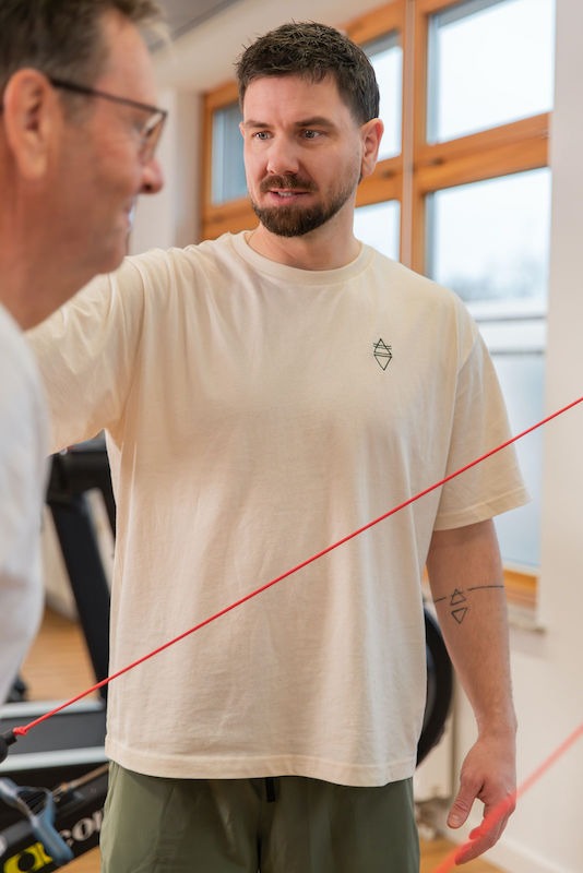 Individuelles-Krafttraining-Move-and-Mind-Harburg Mitja legt Hand an bei einem älteren Patienten, der eine Rückenübung mit einem Widerstandsband macht