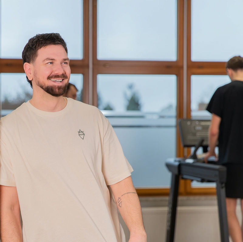 Lächelnder Mitja mit Patienten auf Laufband im Hintergrund