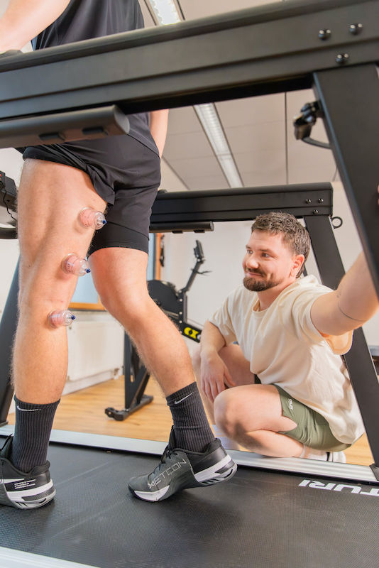 Physiotherapie-Laufband-Move-and-Mind-Harburg Mitja hockt neben einem Laufband und begutachtet einen Patienten, der mit drei Saugnäpfen am hinteren Oberschenkel langsam geht