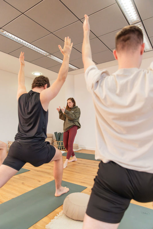 Yogakurse-HH-Harburg-Move-and-Mind Yogakurs: zwei junge Männer von hinten in Kriegerposition, Anne im Hintergrund, gibt Anweisungen