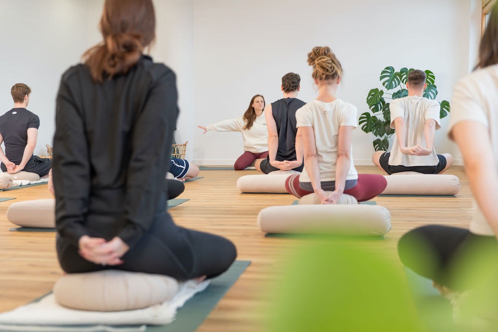 Yogatraining-Harburg-Move-and-Mind Yogagruppe im Schneidersitz mit hinter dem Rücken gestreckten Armen, Yogalehrerin Anne im Hintergrund